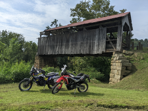 Two dualsport bikes on a clearing in the foreground with a derelict bridge in the background.